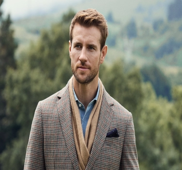 A young man with blonde hair wearing a blue blouse, brown scarve, and a jacket in the summer nature. 