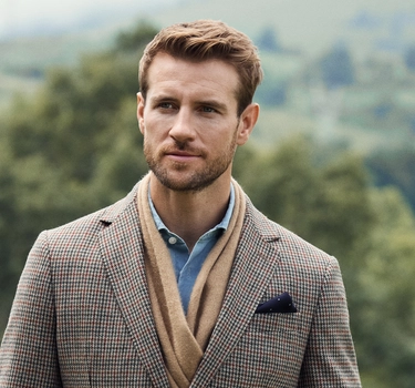 A young man with blonde hair wearing a blue blouse, brown scarve, and a jacket in the summer nature.
