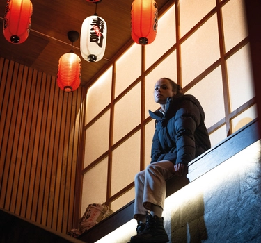 En person sitter på en opphøyd kant i et urbant miljø iført en funksjonell jakke fra SWIMS.