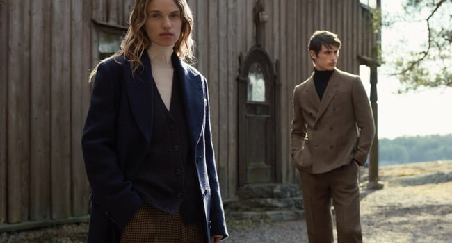 To menn i stilrene kåper og dresser fra Tiger of Sweden foran en rustikk trevegg.