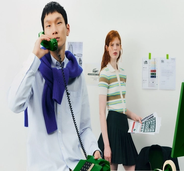 An asian man calling on the phone and a ginger woman at the office. A man wearing a light blue blouse and woman wearing a green striped polo and a black skirt.