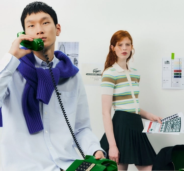 An asian man calling on the phone and a ginger woman at the office. A man wearing a light blue blouse and woman wearing a green striped polo and a black skirt.