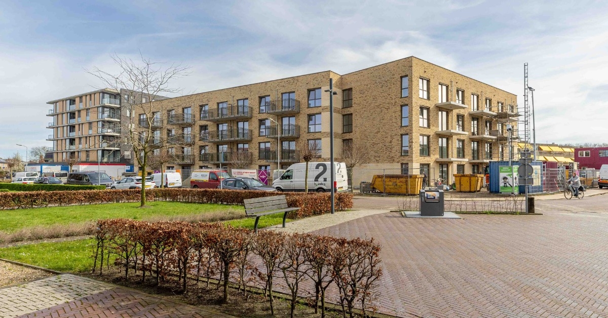 Een modern woongebouw van meerdere verdiepingen met balkons, gebouwd met lichtgekleurde bakstenen en grote ramen. Voor het gebouw staan geparkeerde auto's en busjes, evenals bouwmateriaal zoals steigers en afvalcontainers. Op de voorgrond bevindt zich een groen parkje met gras, gesnoeide heggen, een bankje, een betegeld pad en een lantaarnpaal met het cijfer "2".