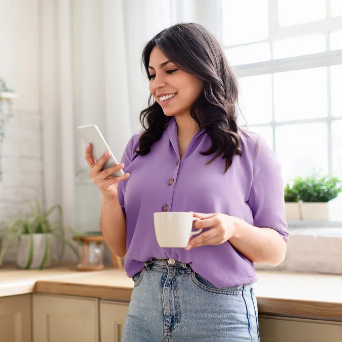 A woman in the kitchen holding a cup and looking at her phone