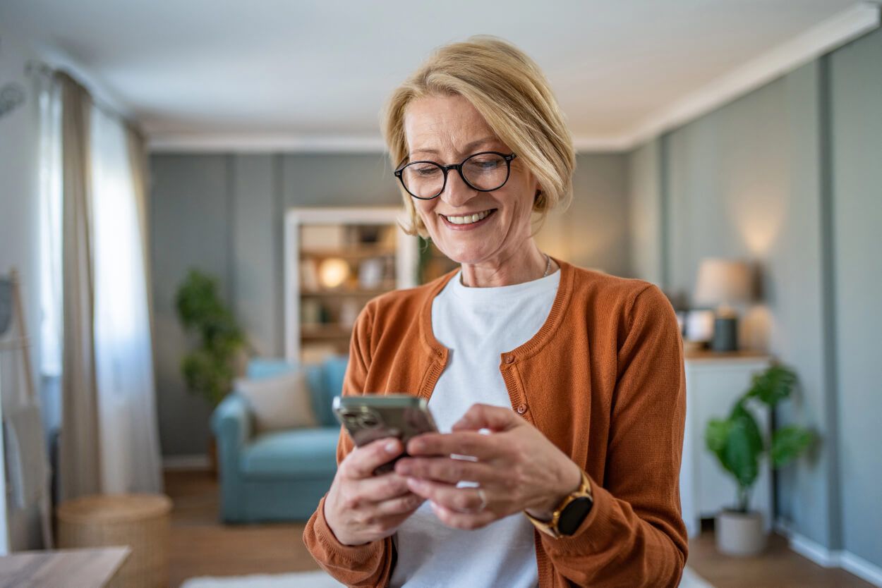 A woman standing in her living room, looking down at her phone and smiling