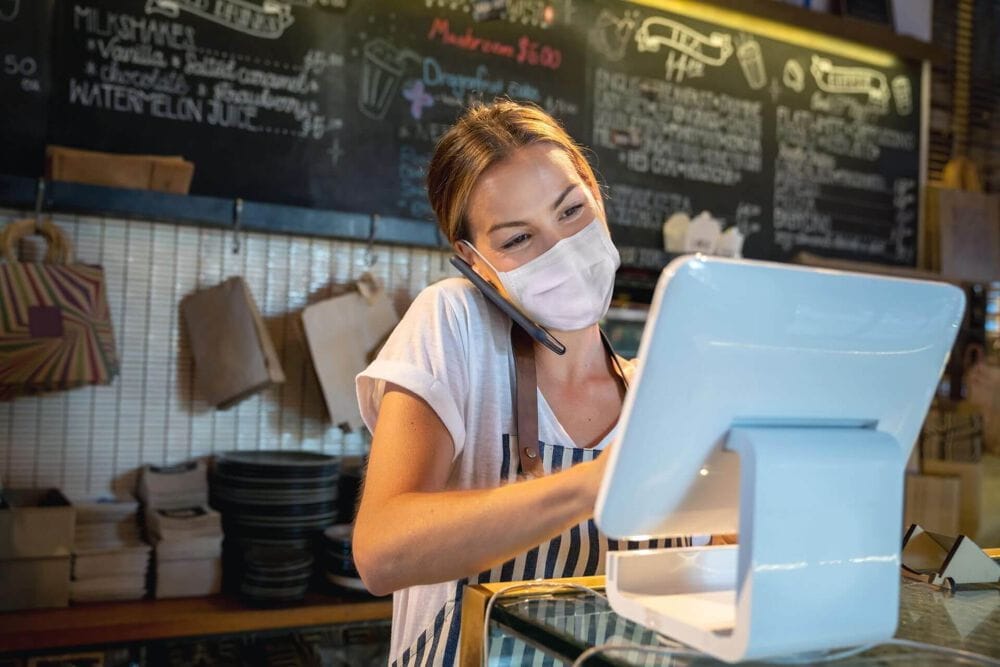 Woman wearing a mask and on the phone working behind the checkout