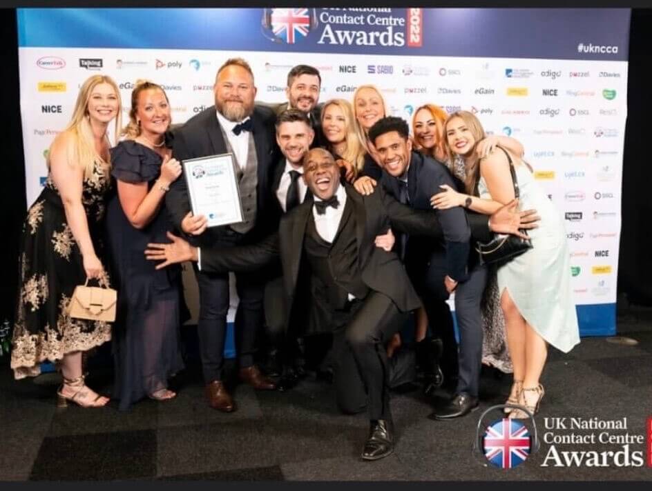 The Atom bank Contact centre team holding an award they won at the UK National Contact Centre awards with Ainsley Harriott crouching and smiling in front of them