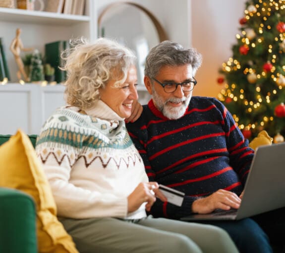 A couple sitting on the sofa smiling whilst looking at their laptop