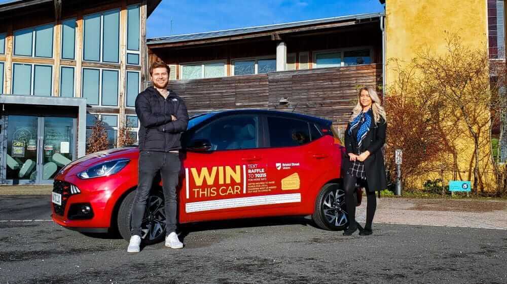 Two people standing in front of a car that is available to win in the raffle