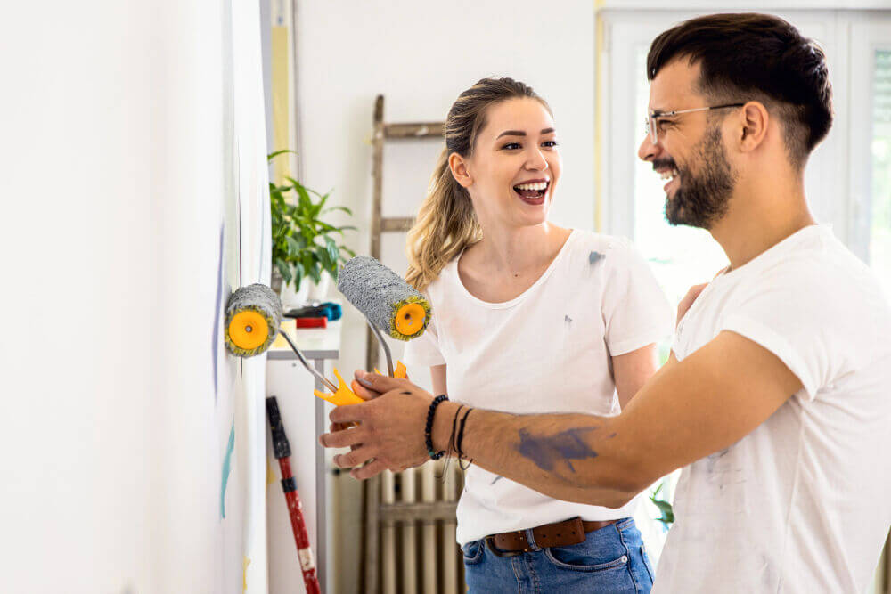 A young couple are smiling as they paint the wall in their home together