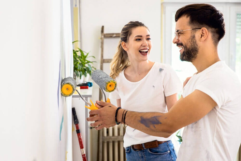 A young couple are smiling as they paint the wall in their home together