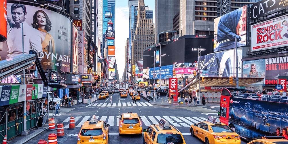 Time square in New York City, there are yellow cabs driving down the street towards the iconic billboards. It is very busy