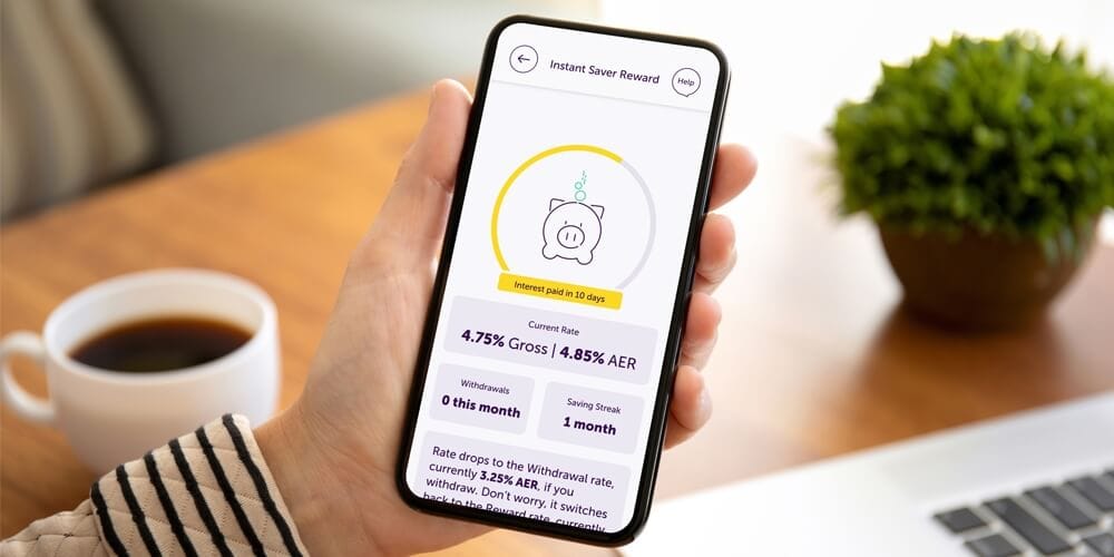 A hand holding a phone showing the instant saver reward rate screen from within the Atom Bank app