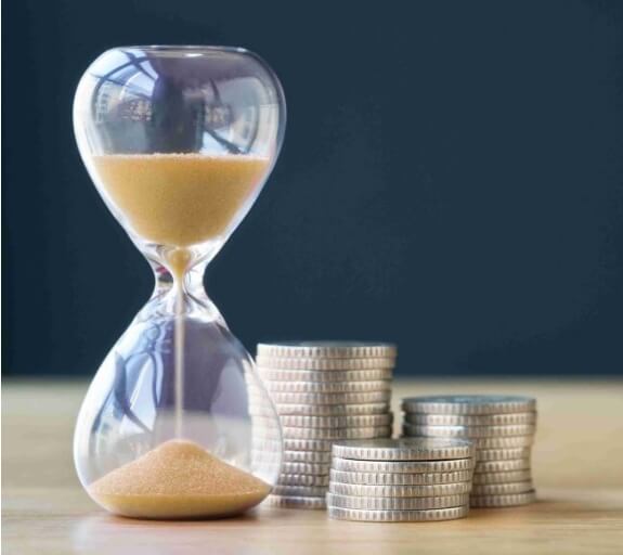 Hour glass with yellow sand falling through it is stood next to a small pile of silver coins