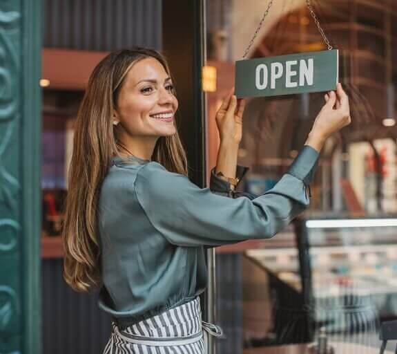 Young woman wearing an apron and smiling is turning a cafe's door sign to Open