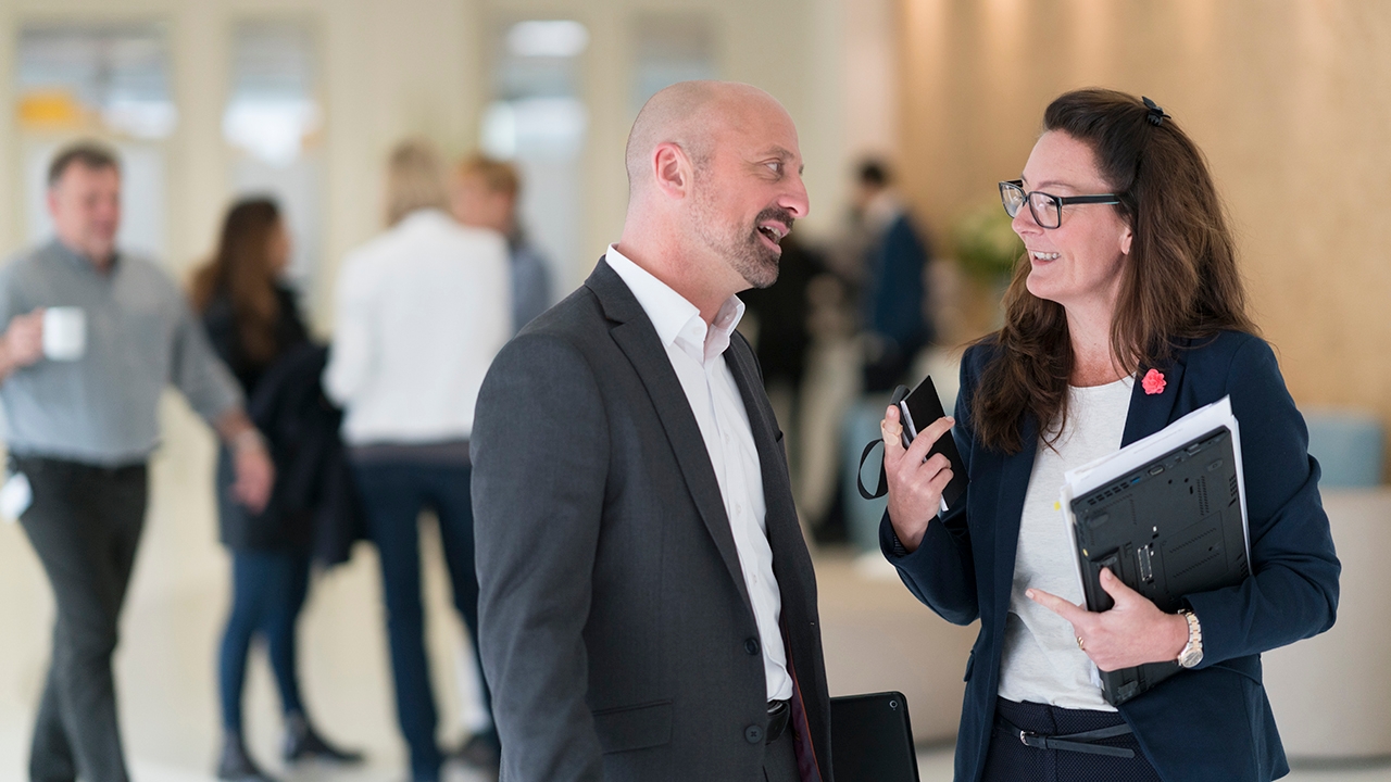 Fotografie eines Mannes und einer Frau in Geschäftsanzügen, die miteinander sprechen. Beide haben einen heiteren Gesichtsausdruck.