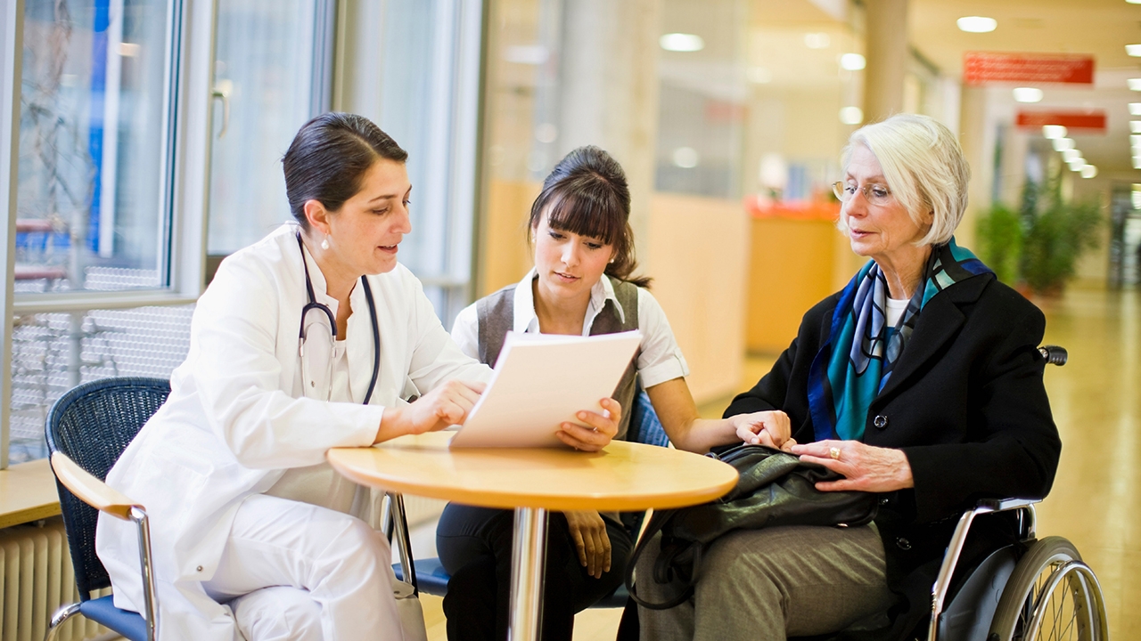 Eine ältere Frau im Rollstuhl sitzt gemeinsam mit einer jungen Frau und einer Ärztin an einem kleinen runden Tisch. Die Ärztin hält ein Dokument in den Händen und liest es vor. Die jüngere Frau hält die Hand der älteren Frau.