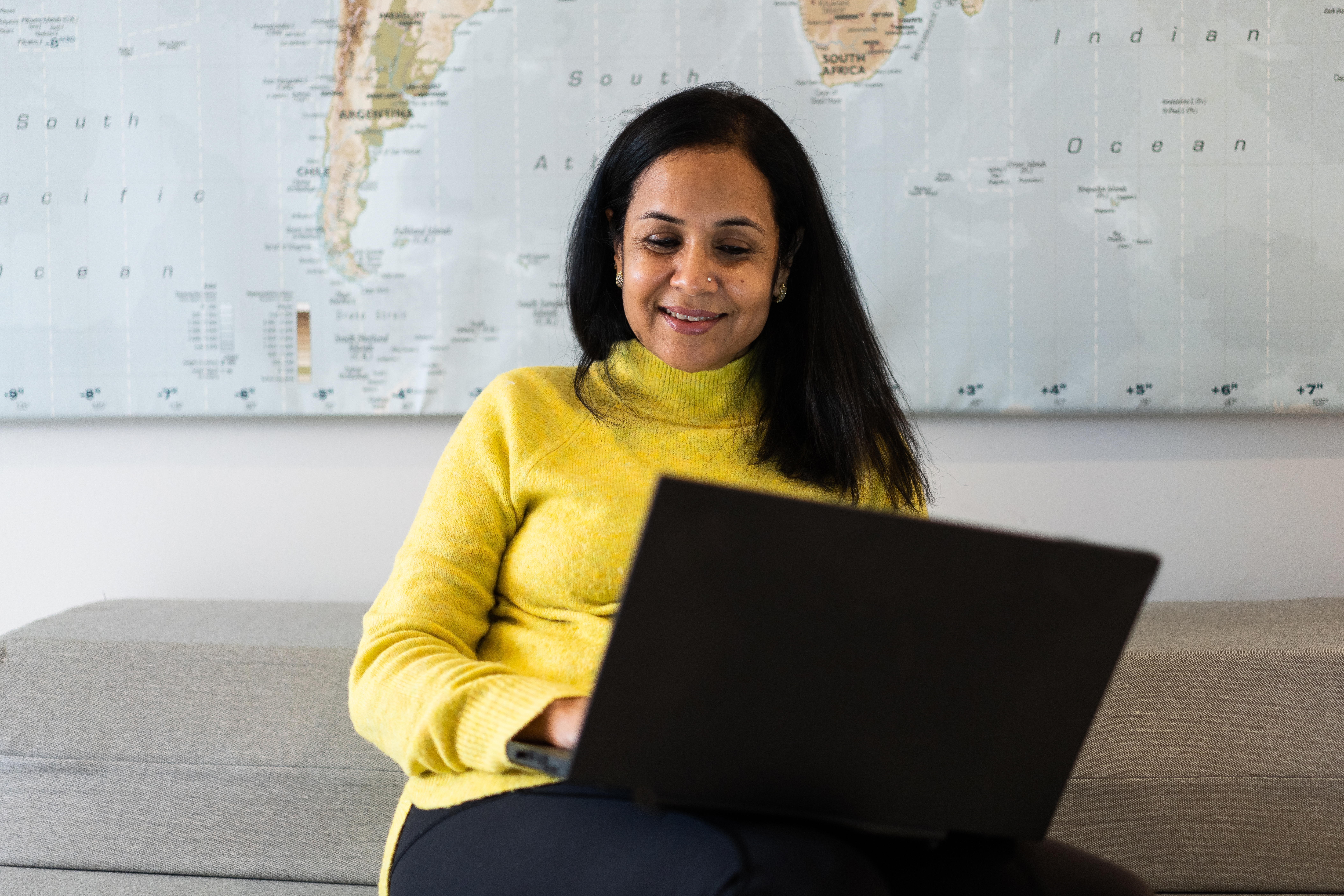 Frontalansicht einer Frau im gelben Pullover, die mit ihrem Laptop auf einem Sofa sitzt.