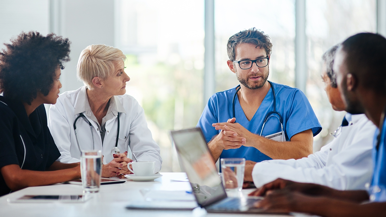 Fotografie von fünf Personen, die an einem Konferenztisch sitzen. Alle tragen medizinische Kittel mit einem Stethoskop um den Hals. Der Mann in der Mitte spricht, während die anderen vier Personen am Tisch ihn anschauen.