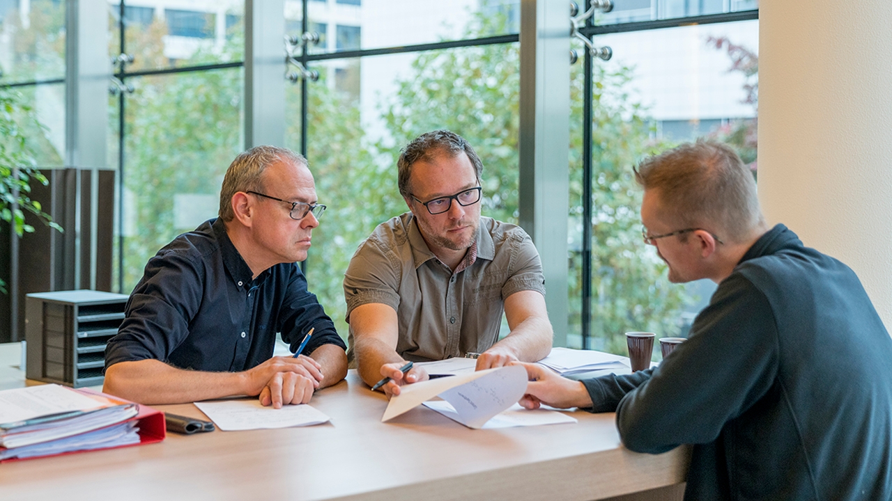 Drei Personen sitzen zusammen an einem Tisch in einem hellen, lichtdurchfluteten Raum. In der Tischmitte liegen Dokumente, auf die eine der Personen zeigt, während sie gleichzeitig Blickkontakt mit der Person gegenüber sucht.