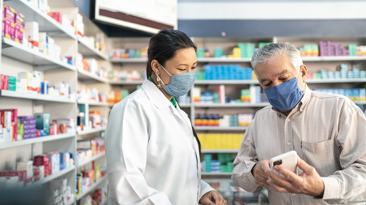 Ein älterer Herr mit Mundschutz zeigt einer Apothekerin ein Bild auf seinem Smartphone. Im Hintergrund befinden sich Regale mit Arzneimitteln.
