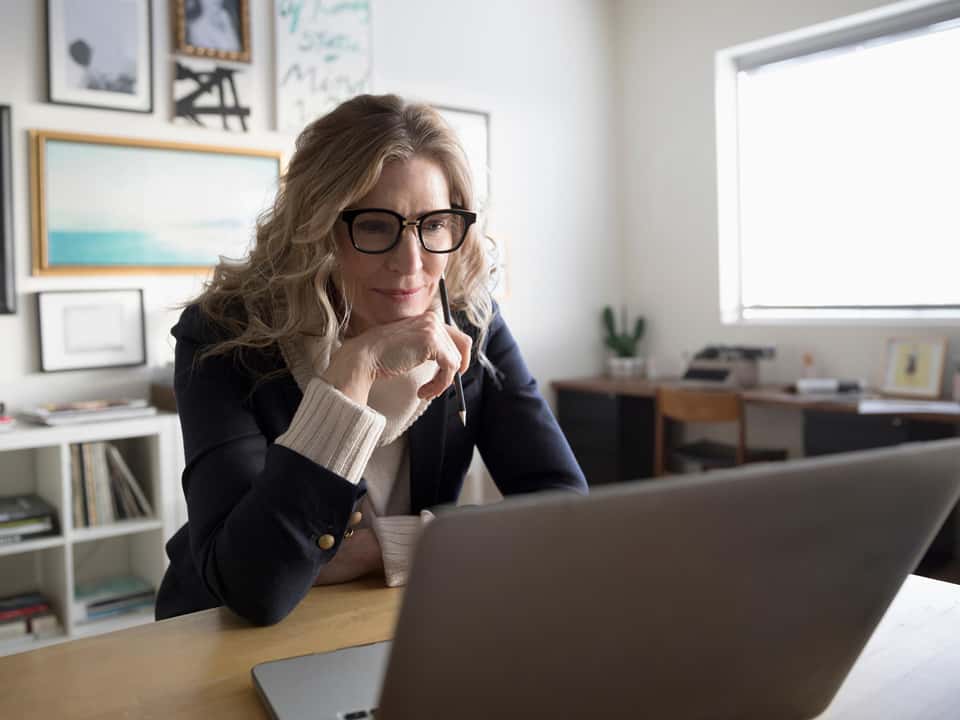 Frau mit Brille arbeitet konzentriert an einem Laptop im Homeoffice Eine Frau mittleren Alters mit Brille, sitzt in einem stilvoll eingerichteten Homeoffice und schaut konzentriert auf den Bildschirm ihre Laptops. Im Hintergrund sind Bücherregale, Bilder und ein Schreibtisch mit Pflanzen und Schreibmaschine zu sehen.