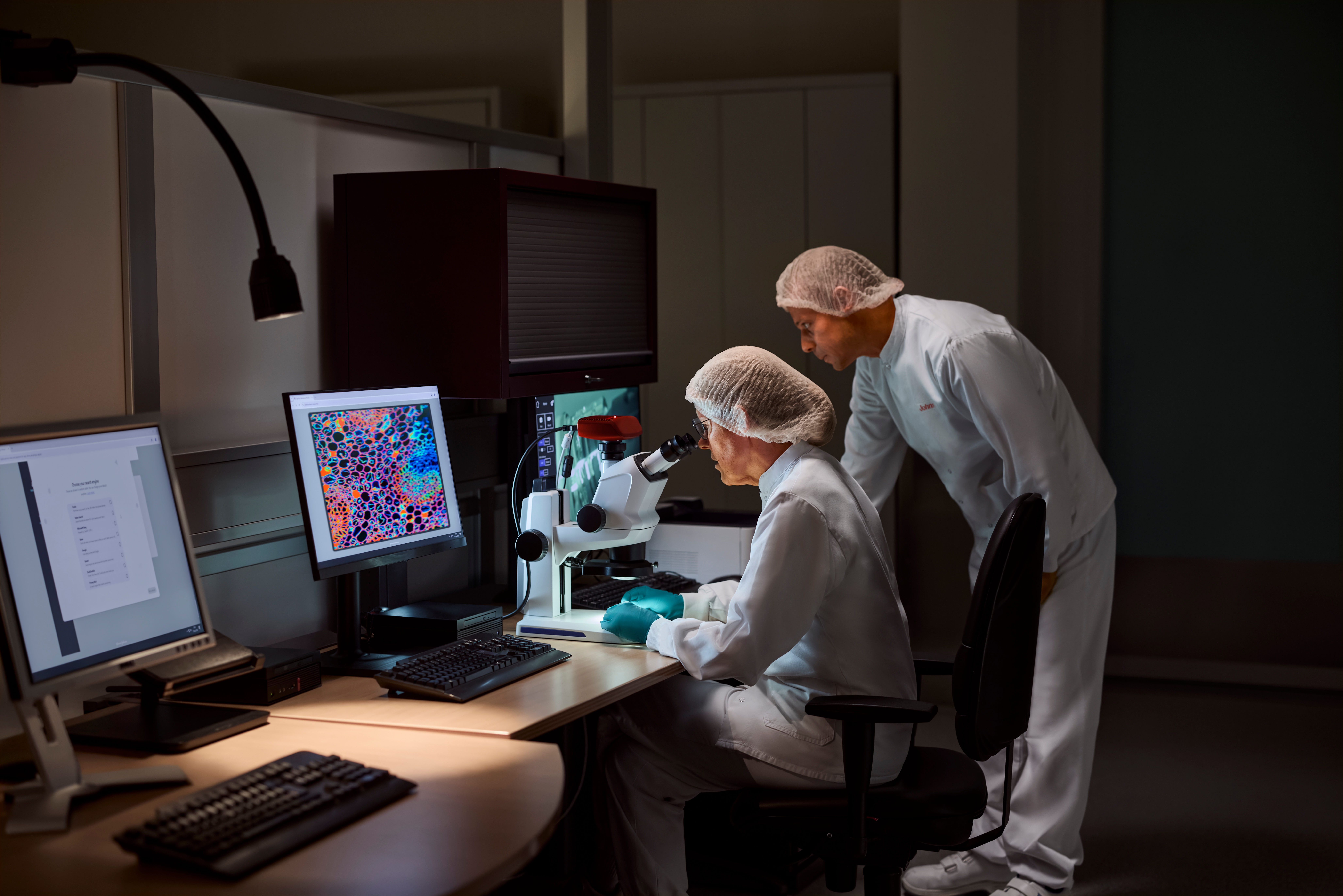 Ein Mann und eine Frau arbeiten in einem abgedunkelten Raum vor medizinischen Geräten. Beide tragen Kittel und Haarnetz. Die Frau schaut in ein Mikroskop, während der Mann einen Bildschirmmonitor anschaut.