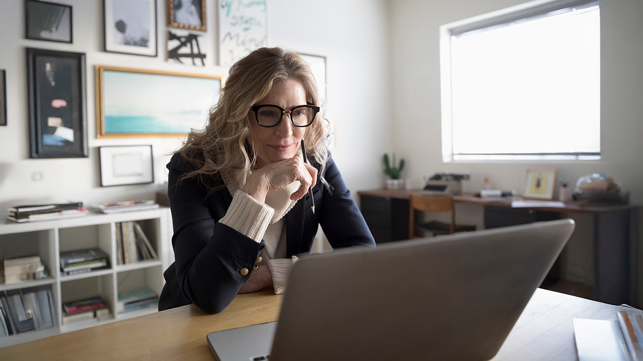 Fotografie einer Frau mit Brille und blonden Haaren, die auf den Bildschirm eines Laptops schaut. Dabei stützt sie ihren Kopf auf ihre rechte Hand.
