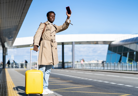 A young man hails a taxi after a trip where his belongings were protected by travel insurance. A young man with a phone in his hand and yellow luggage hails a taxi at the airport.