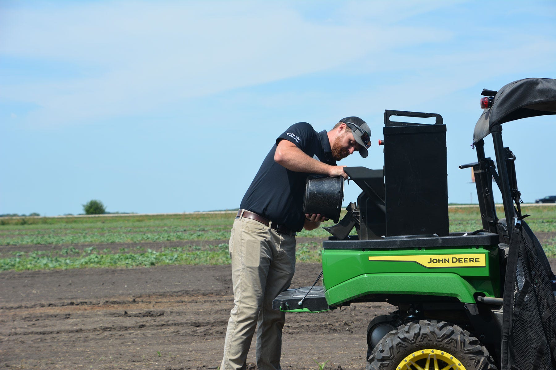 man pours something from a bucket into the back of a John Deere Gator out in the field