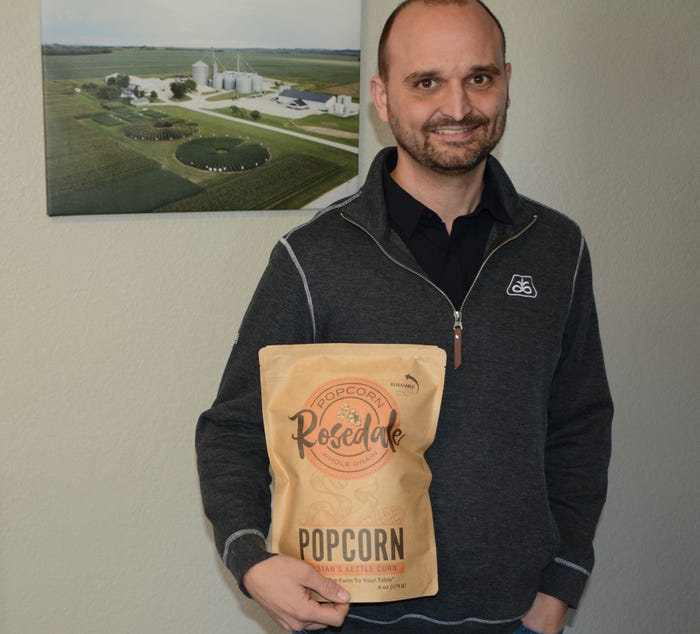 Wes Wagler holds a bag of Rosedale Popcorn while standing in front of a farmstead photo