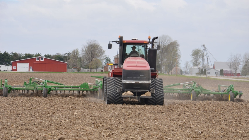 A tractor pulling a tillage implement in a no-till cornfield with a farmstead in the background 