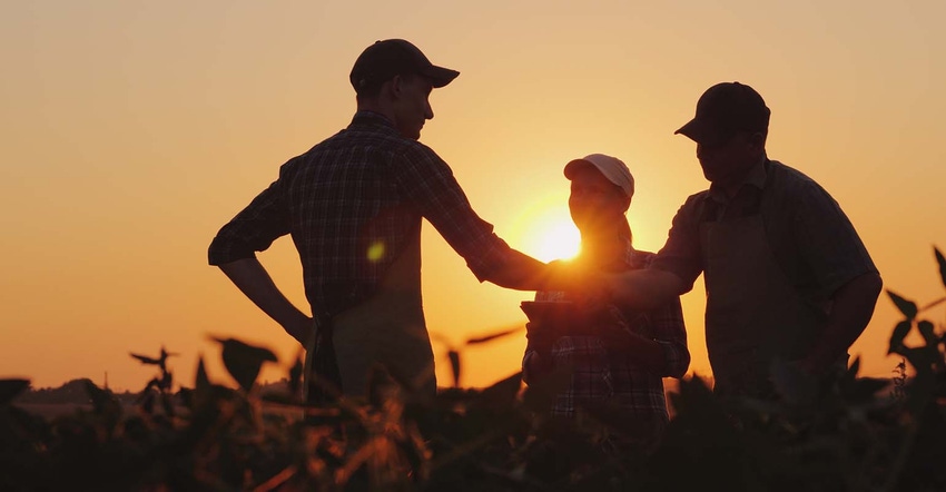 Three people standing in a farm field at sunset shaking hands.