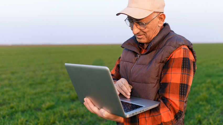 Man standing in field holding a laptop with one hand and typing with the other hand.