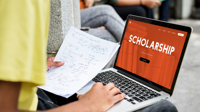 student holding papers while looking at the words scholarship on computer screen