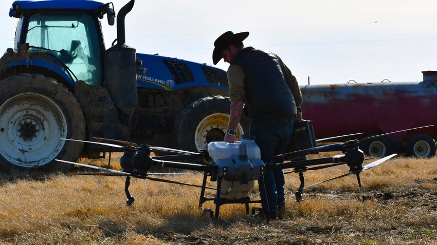 Man adjusting an agricultural spray drone on the turnrow of a field with a tractor in the background.