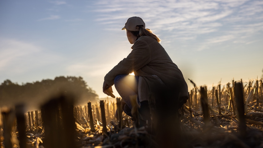 Farmer kneeling in corn stubble with sun in background