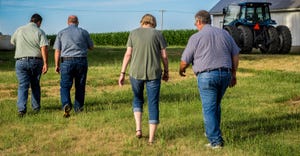 a woman and three men walking away from the camera toward a tractor and a cornfield