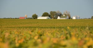 A landscape of a field with a barn and silos in the distance near the tree line
