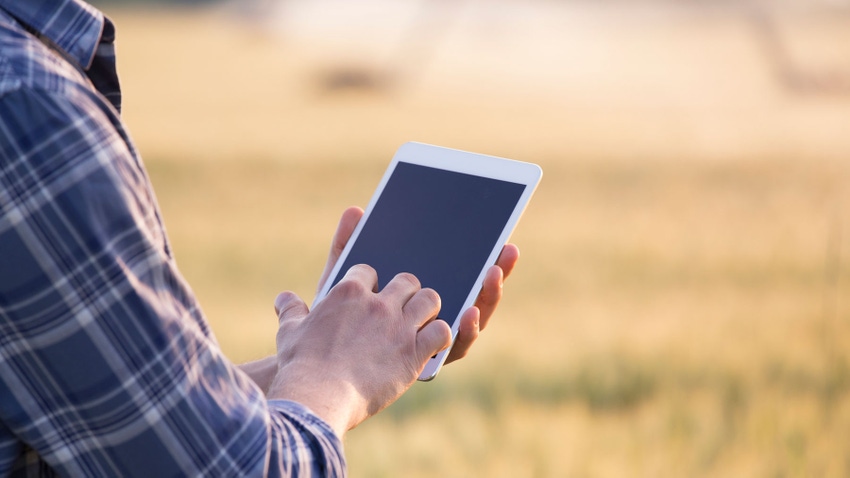 Close up of a person's hands holding a smart tablet, wearing a plaid collared shirt, with a field in the background.