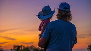 A young man holding a toddler wearing a cowboy hat in his arms as they look toward a sunset