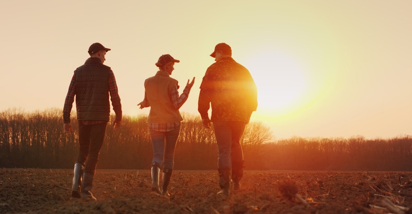 three farmers walking across plowed field towards the setting sun
