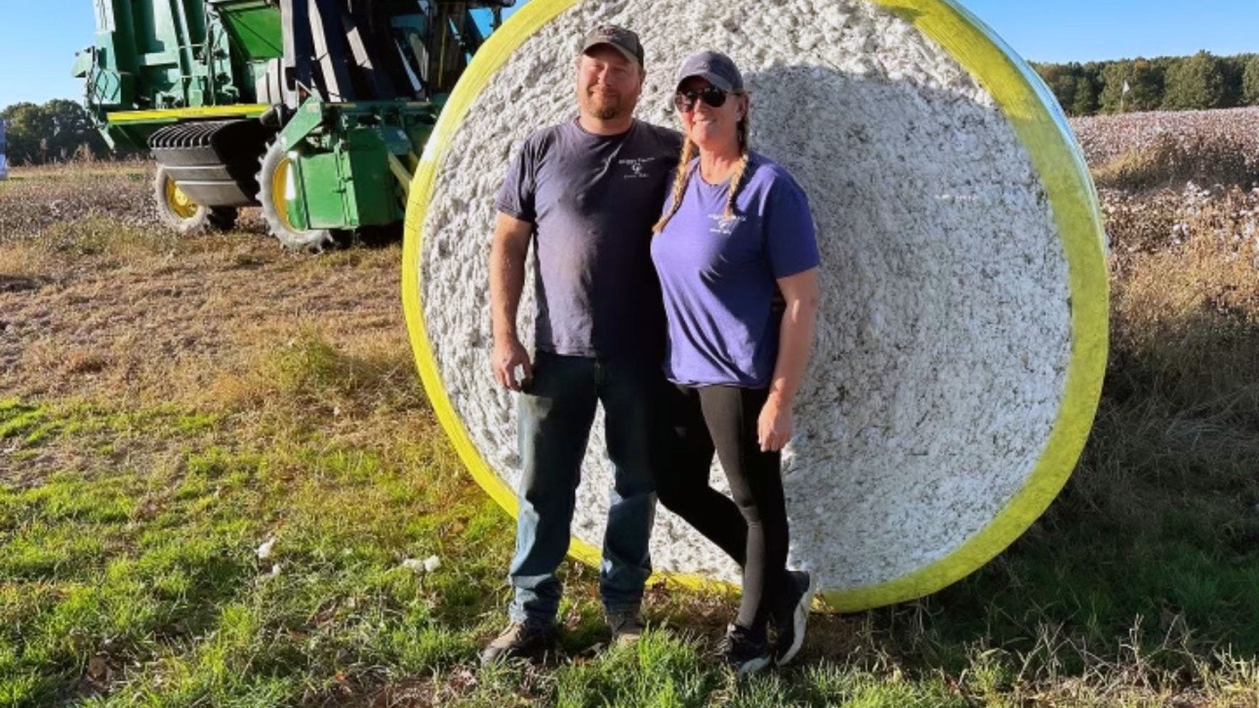 Man and woman farmers standing together in front of a cotton bale wrapped in yellow wrap with cotton picker in background.