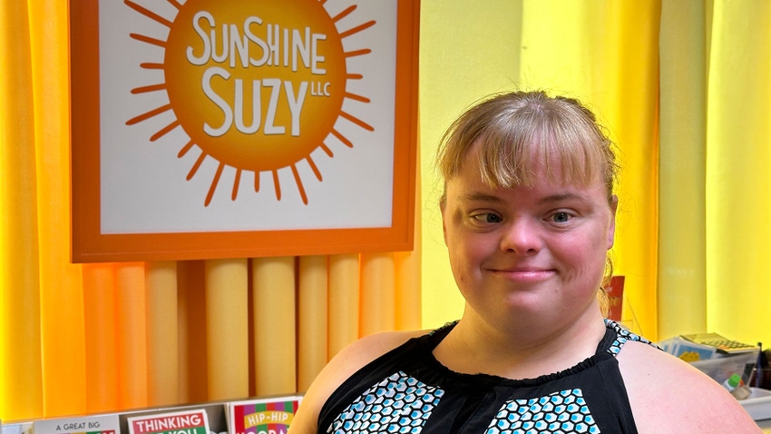 Suzy Sukalski smiles next to a picture of the Sunshine Suzy logo