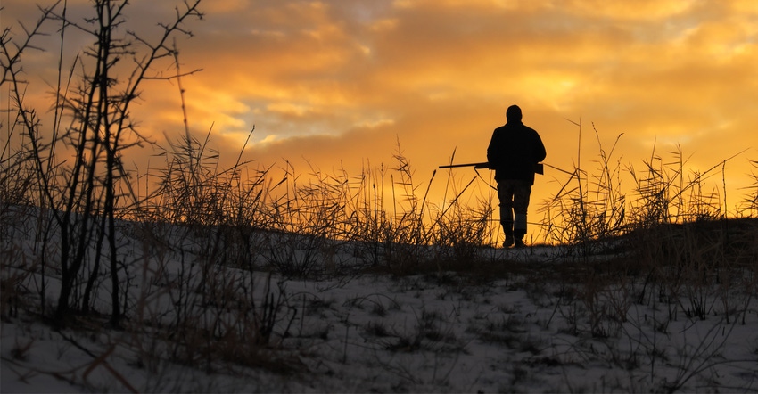sillouette of man with gun walking on snowy ground with sun setting
