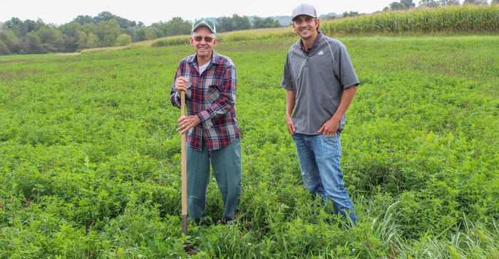 Two men standing in a field while one of them holds a shovel as they smile for a photograph