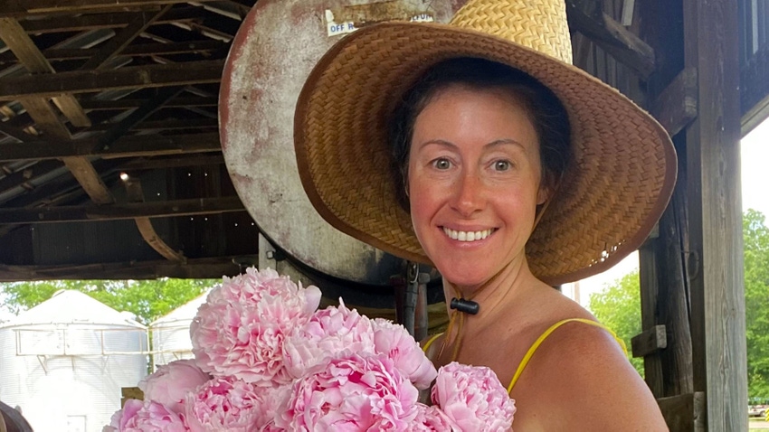 Woman farmer wearing large straw sunhat holding an armful of pink peonies.