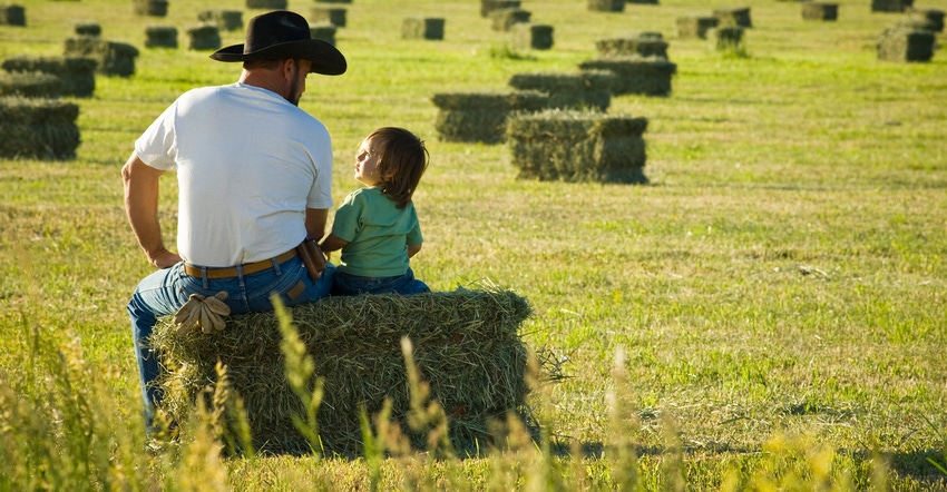 father and child sitting on bale of hay looking out over field