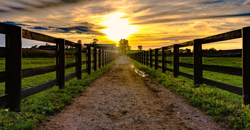 fence-lined dirt road leading to barn with sunset in background