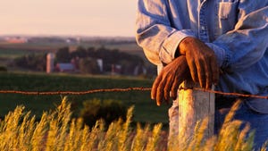 Torso of farmer leaning on fence post with farm in background